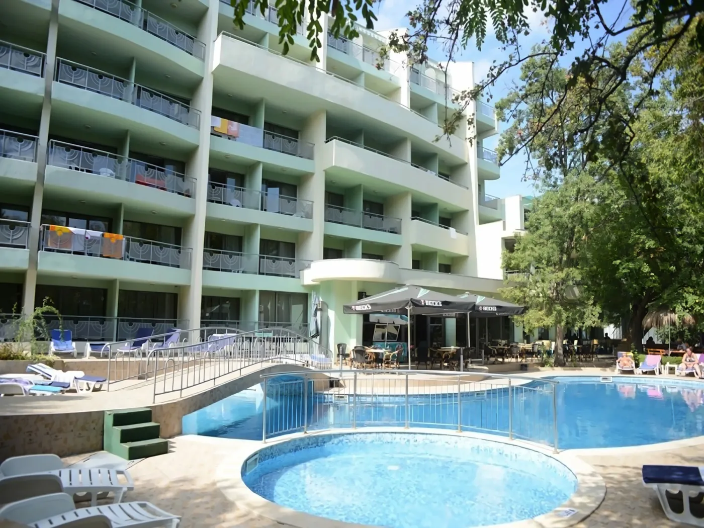 Zona de piscină cu șezlonguri și o clădire verde în fundal. - Zona de piscină cu șezlonguri și o clădire verde în fundal.